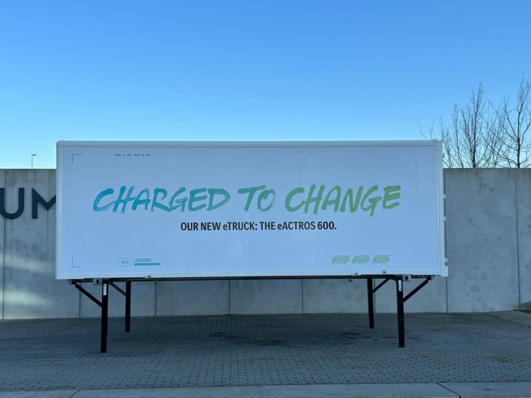 Ein großer weißer Container mit grünem Text "Charged to Change" vor einer Betonwand.