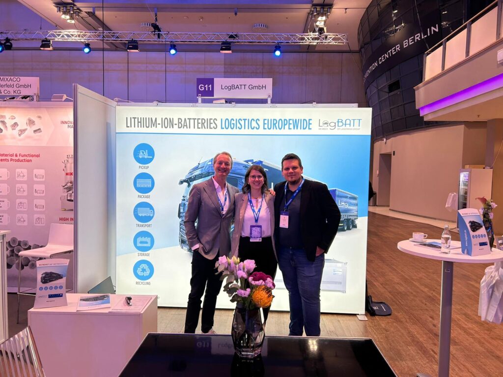 Three people smiling at an exhibition stand with blue advertising for lithium-ion batteries.
