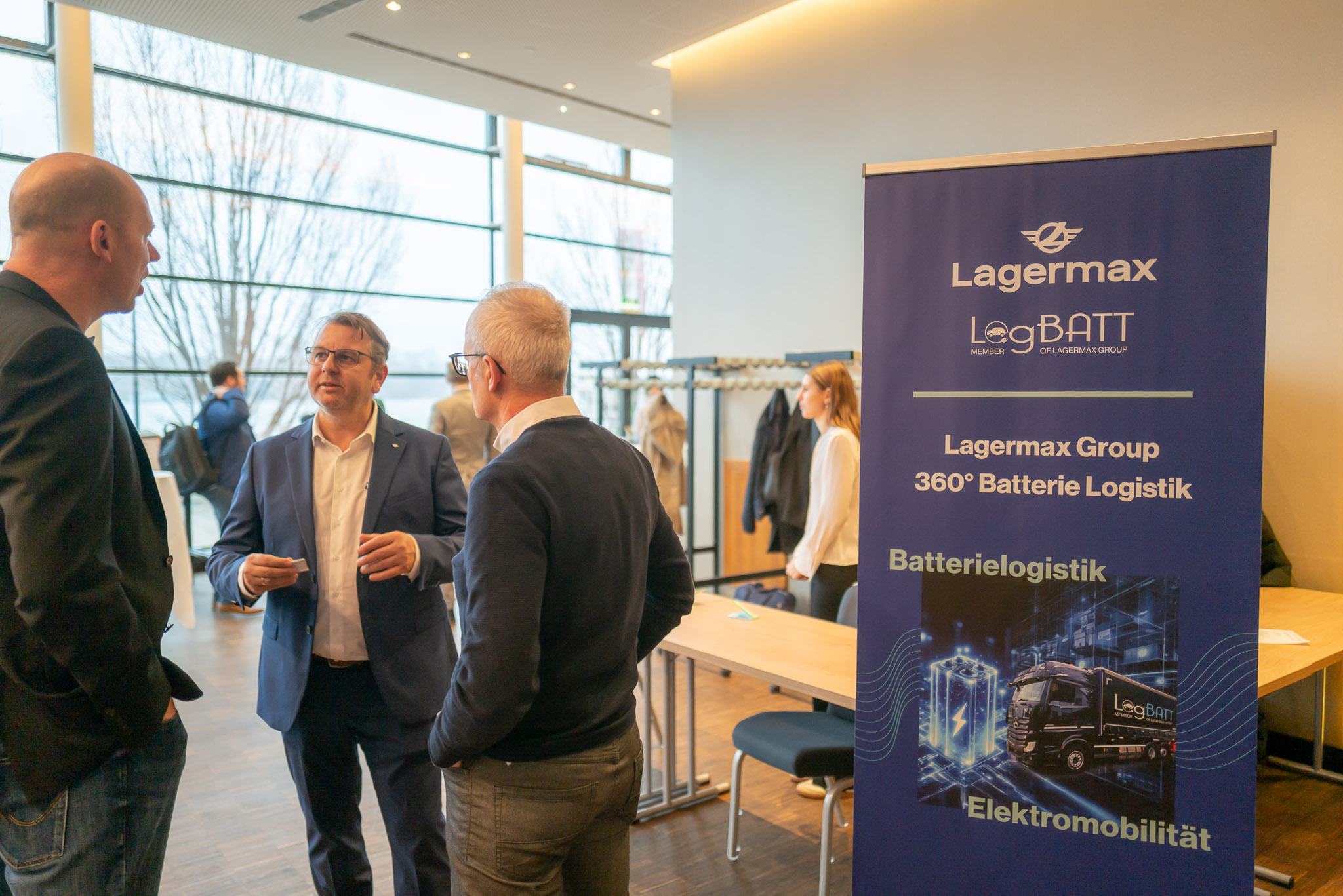 Drei Männer im Gespräch neben einem blauen Banner für Batterie-Logistik in einem modernen Raum.
