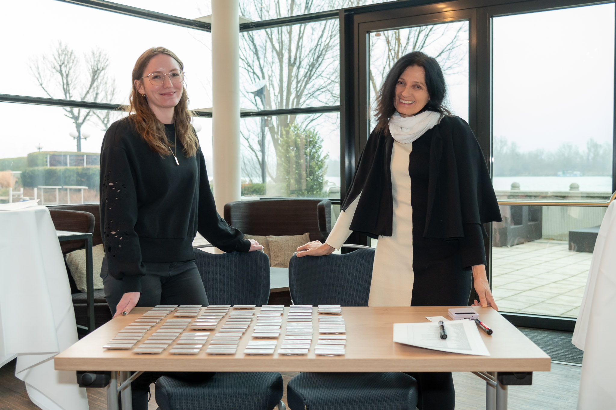 Zwei lächelnde Frauen in Schwarz und Weiß an einem Tisch mit Namensschildern vor einer Fensterfront.