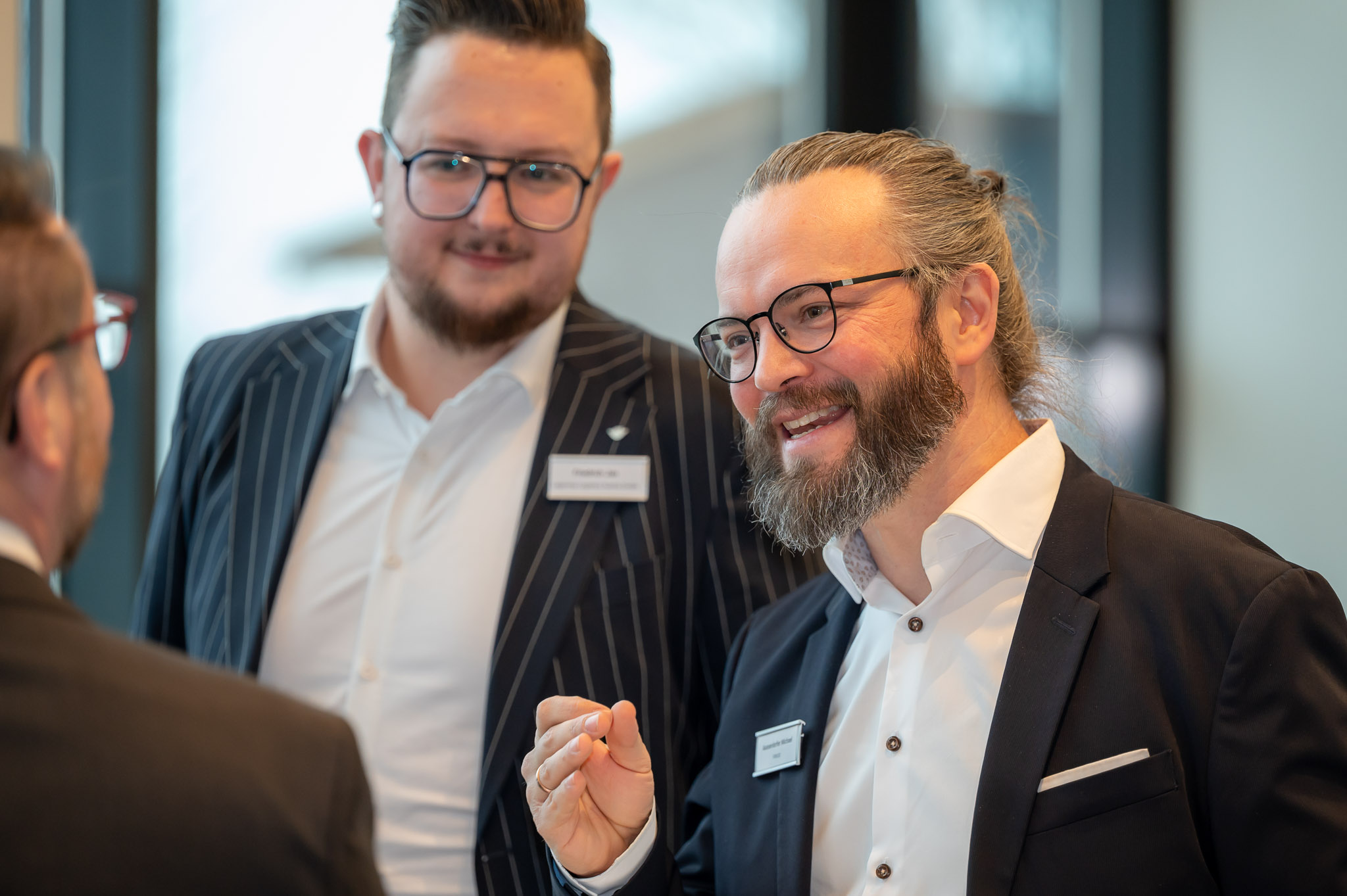 Zwei Männer in Anzügen, freundlich lächelnd, mit Brillen und Namensschildern, im Gespräch.