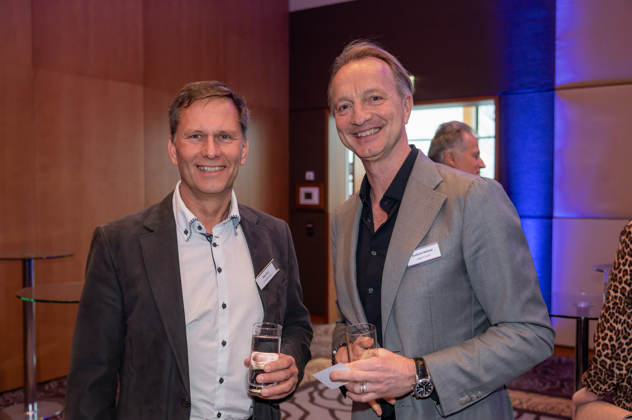 Zwei lächelnde Männer in Anzügen mit Getränken in der Hand, in einem modernen Raum.