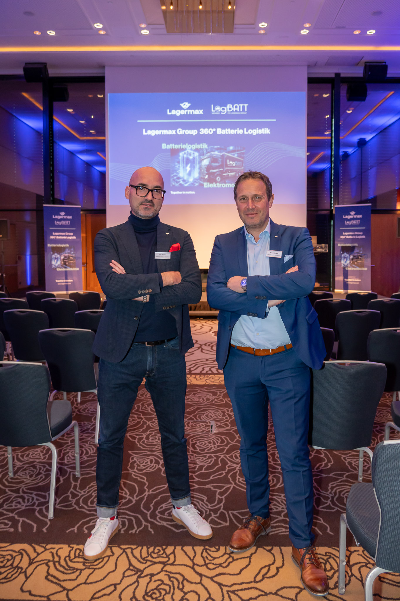 Zwei Männer in Anzügen lächeln, in einem Konferenzraum mit blauen Lichtern und Präsentationsfolie.