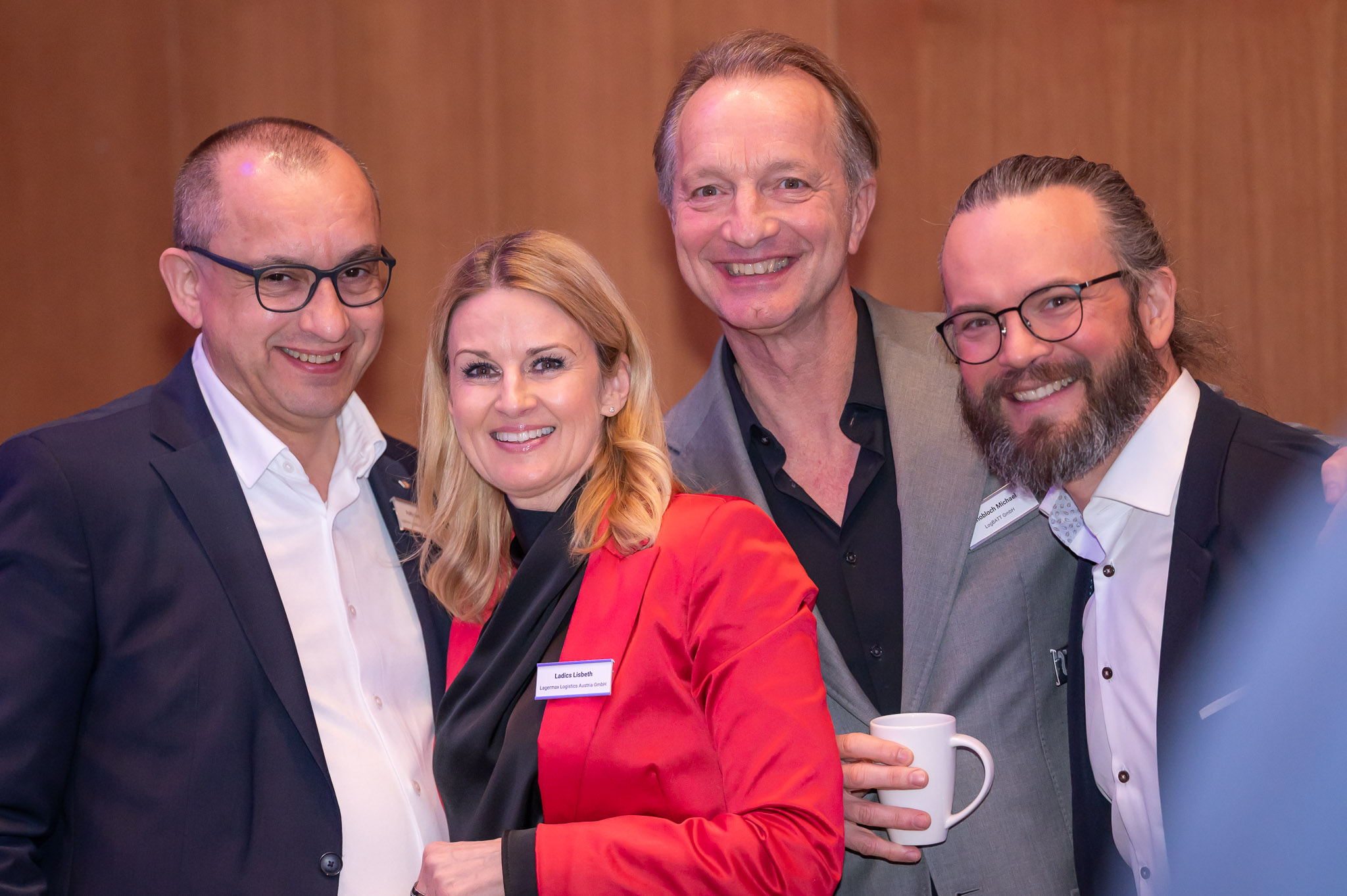 Vier Personen lächeln, eine Frau trägt rot, zwei Männer tragen Anzüge, einer hält eine Tasse.