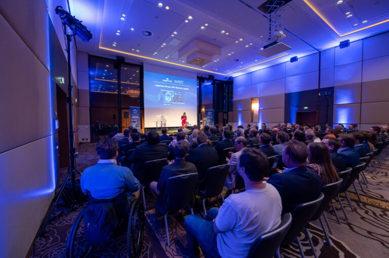 Ein Vortragender in rotem Anzug spricht vor einem Publikum in einem modernen Konferenzraum.