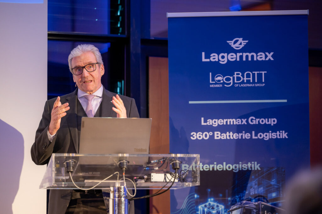 Ein älterer Mann spricht am Rednerpult; trägt Brille und Anzug neben Logistik-Banner.