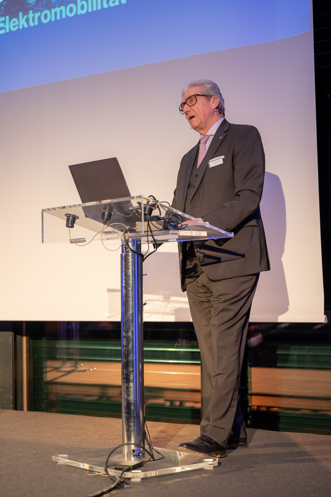 Ein älterer Herr spricht bei einer Präsentation, trägt Anzug und Brille, wirkt ernst.