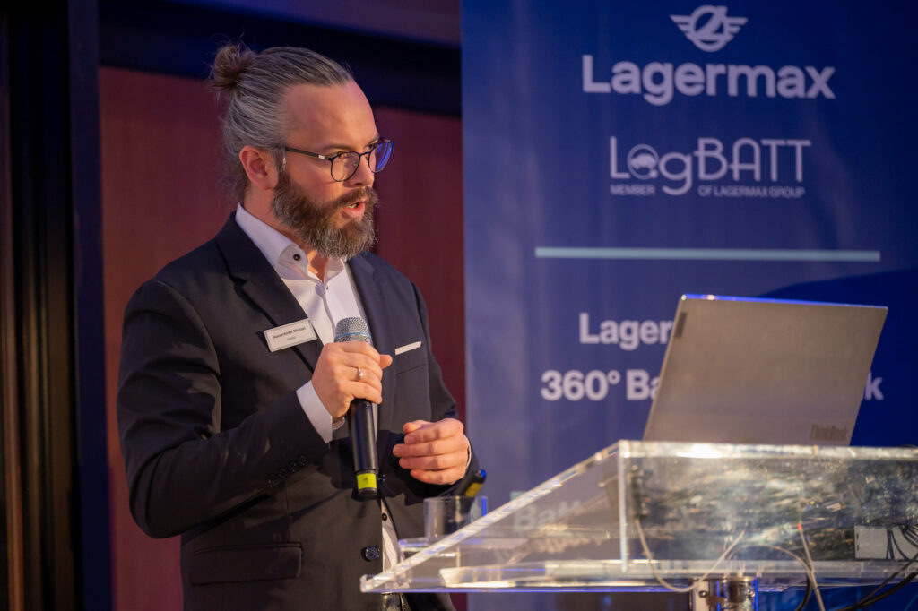 Mann mit Brille und Bart spricht am Pult; formeller Anzug, Mikrofon in der Hand, neutraler Ausdruck.