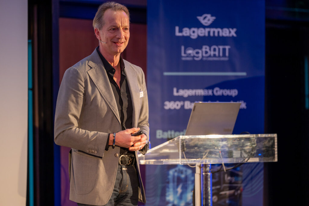 Mann in grauem Sakko spricht lächelnd bei Präsentation, blaues Lagermax-Banner im Hintergrund.