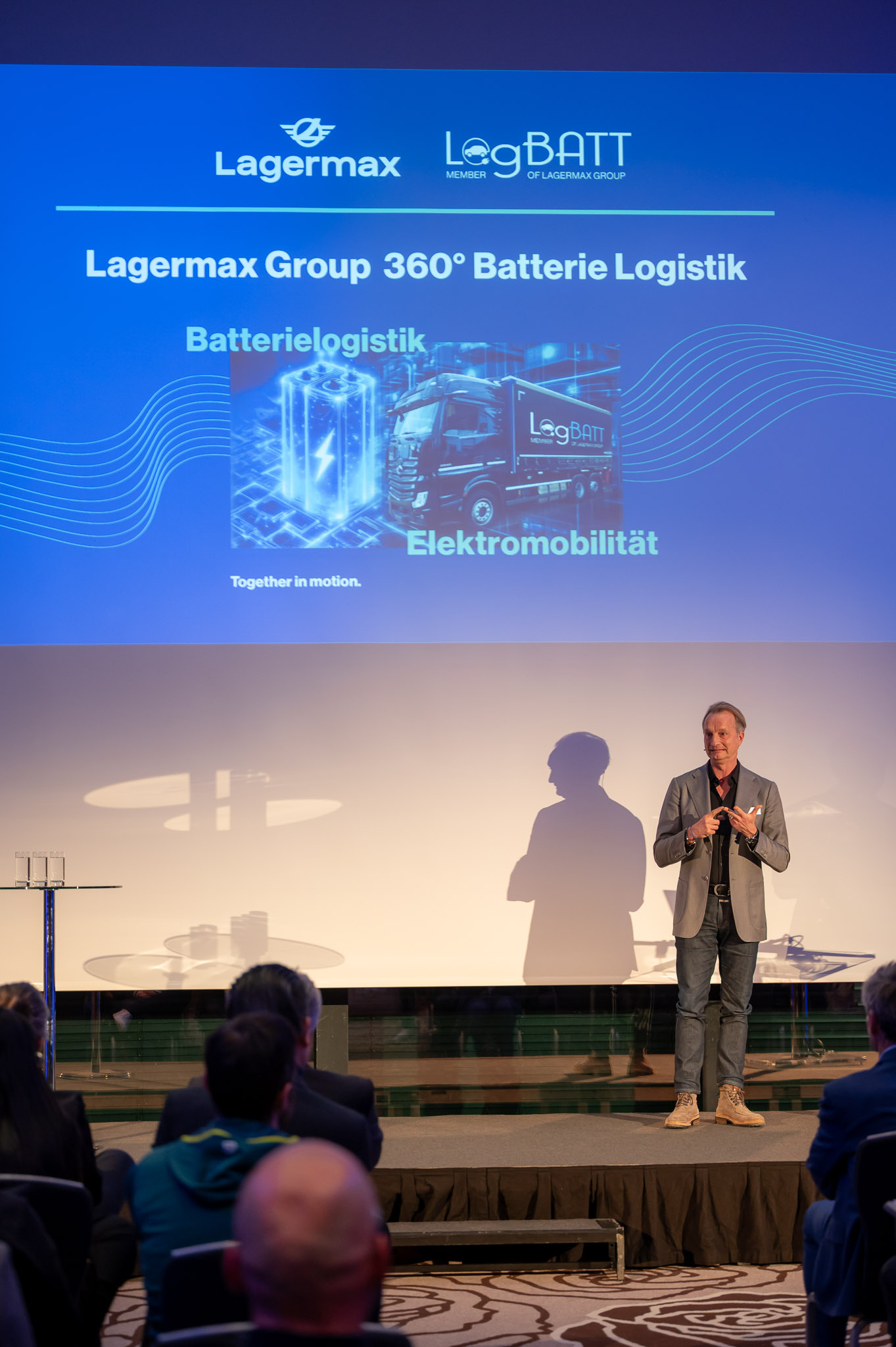 Ein Mann in Jeans und Blazer hält eine Präsentation auf einer Bühne mit blauer Projektionswand.