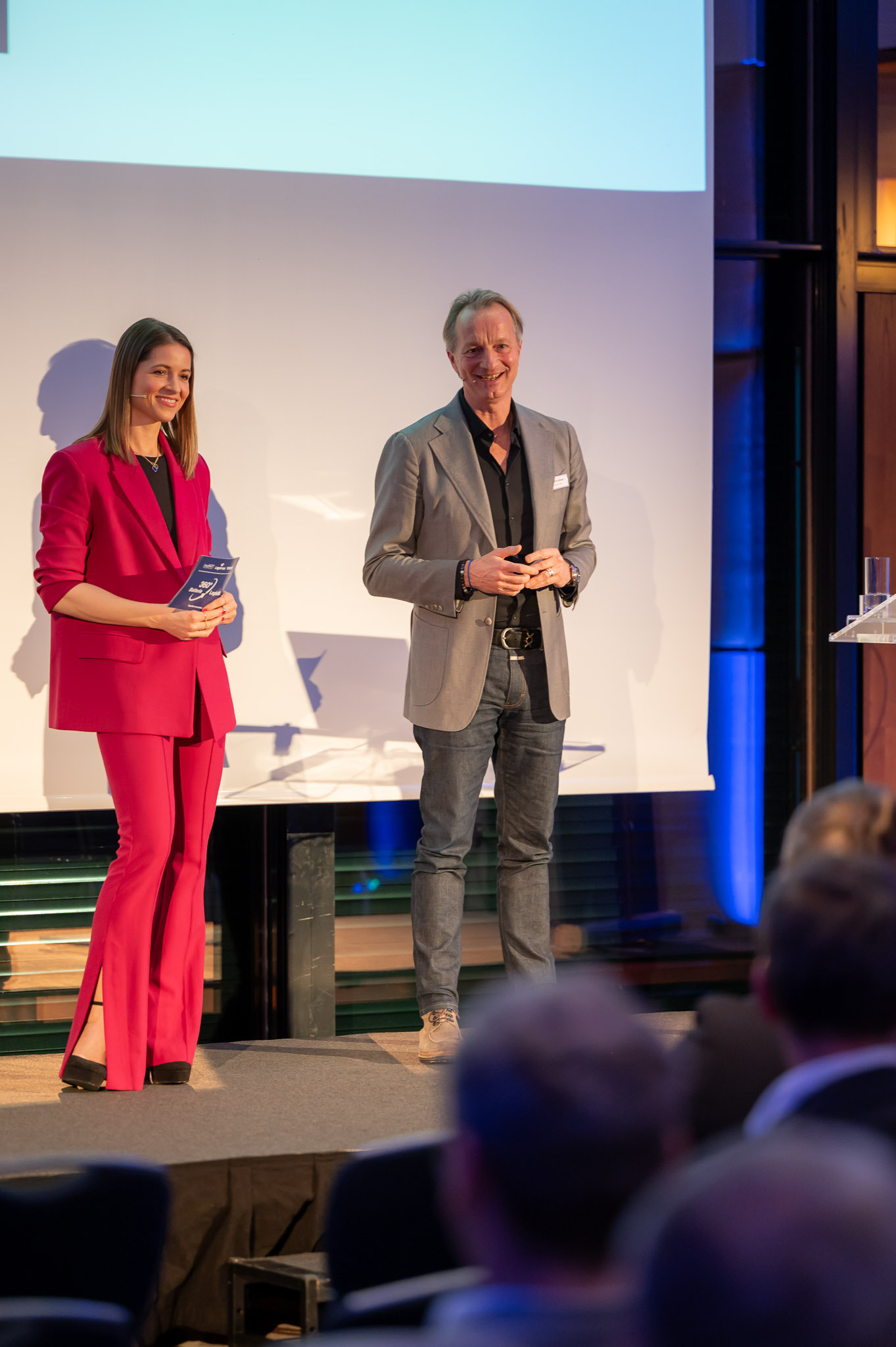 Eine Frau in einem pinken Anzug und ein Mann in einem grauen Blazer auf einer Bühne lächelnd.