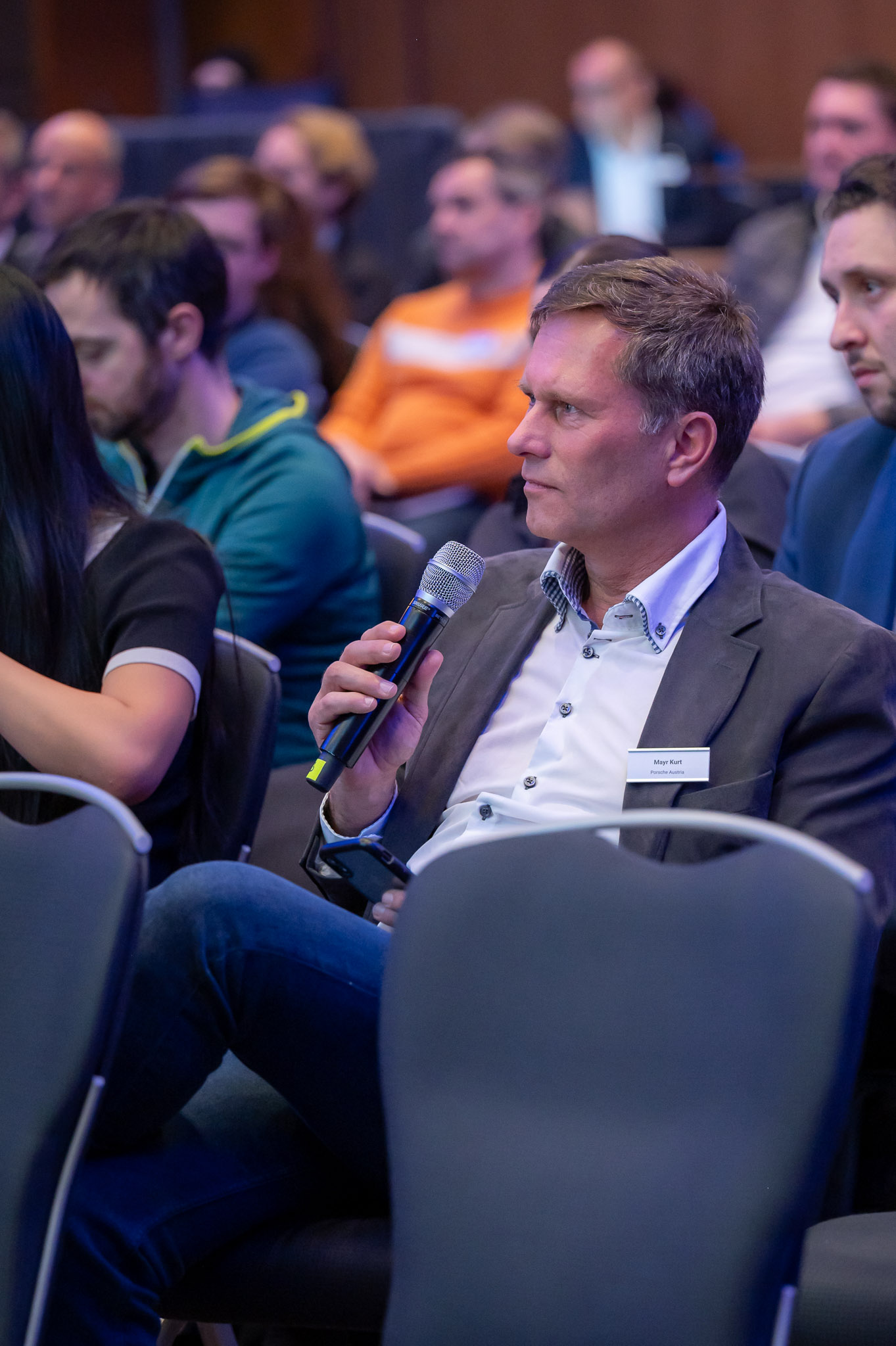 Ein Mann im Anzug spricht ins Mikrofon, sitzt in einer Menschenmenge, ernsthaft und aufmerksam.