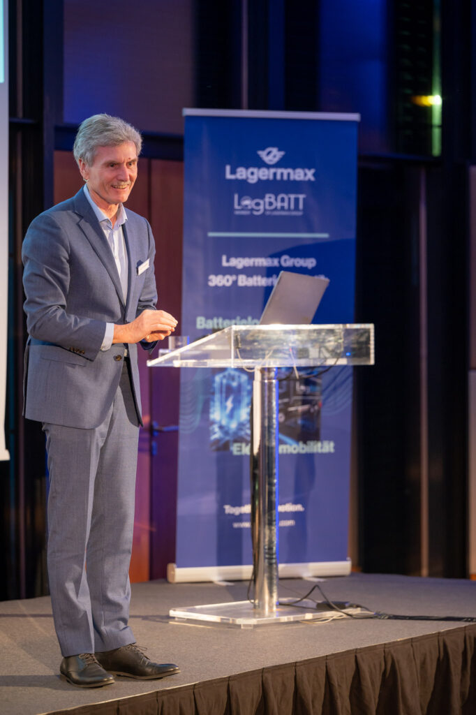 Ein lächelnder Mann im blauen Anzug spricht auf einer Bühne mit einem Laptop.