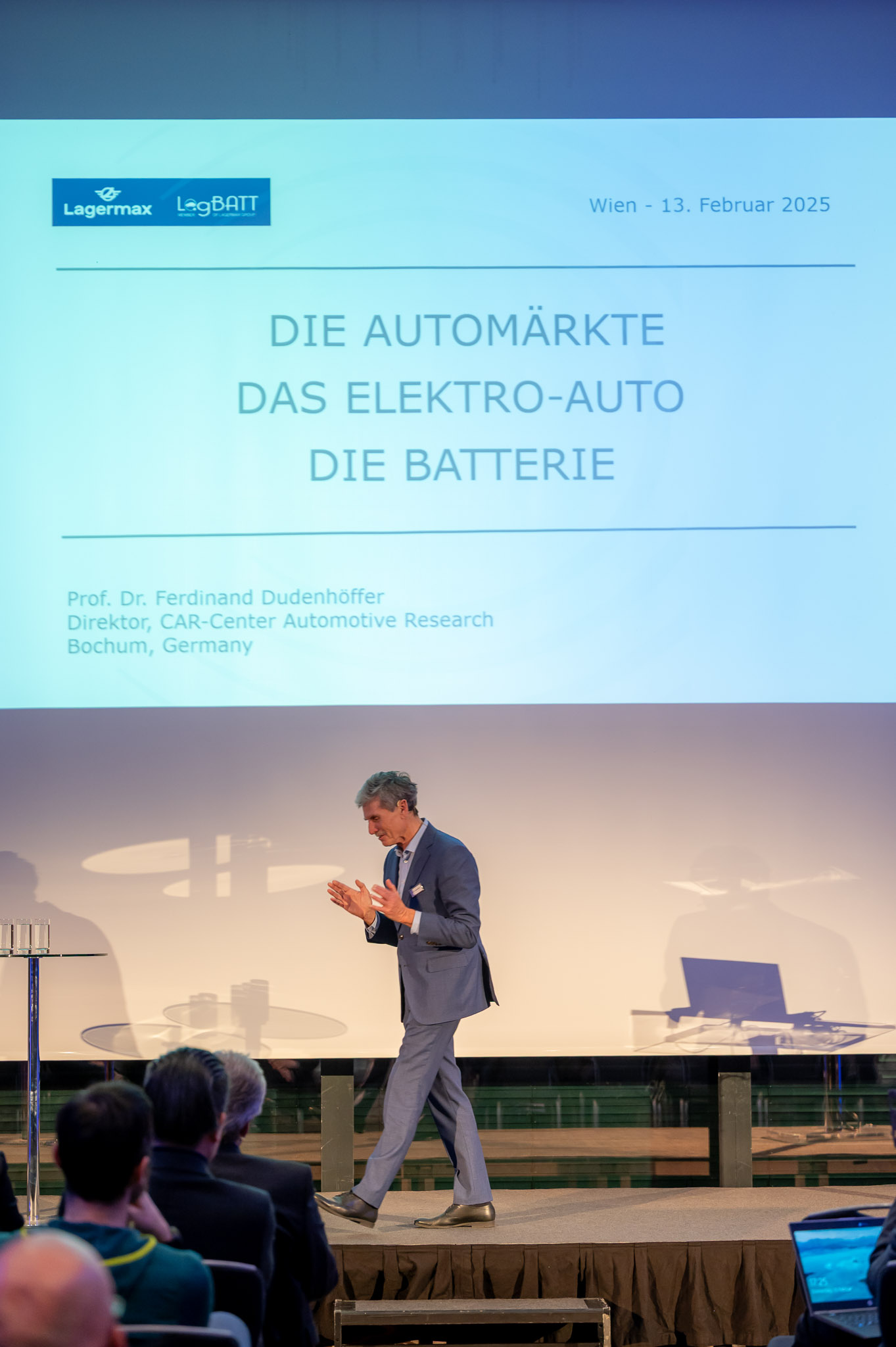 Ein Mann im Anzug spricht vor einem Publikum zu einer Präsentation über Elektroautos und Batterien.