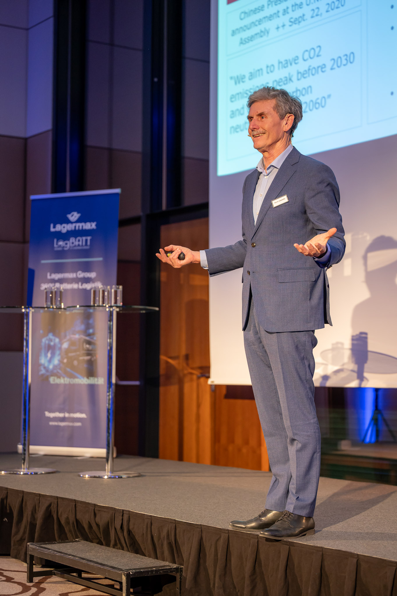 Mann im blauen Anzug lächelt auf Bühne; hält Vortrag vor Projektionsleinwand.