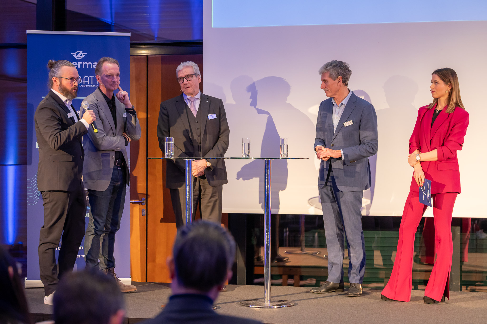 Fünf Personen in formeller Kleidung bei einem Podiumsgespräch; zwei sprechen, andere hören zu.