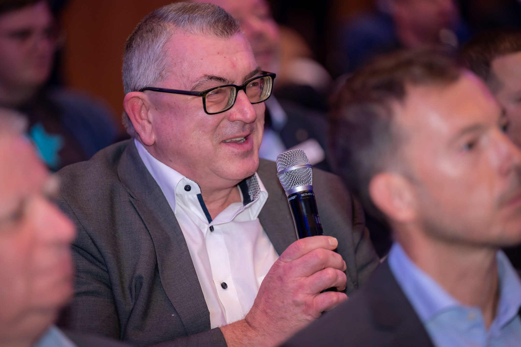 Älterer Herr mit grauem Anzug lächelt, Mikrofon in der Hand, trägt Brille und weißes Hemd.