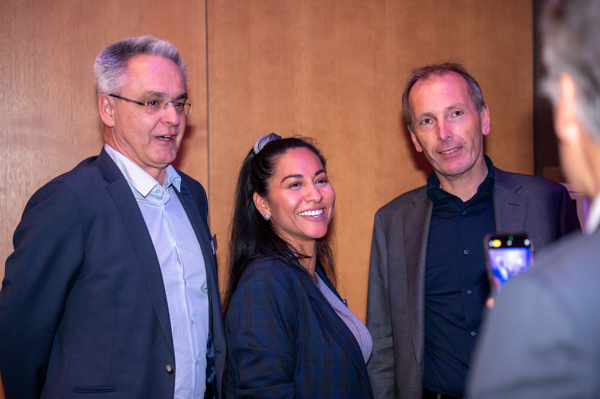 Eine lächelnde Frau zwischen zwei Männern in Anzügen, hölzerner Hintergrund, alle schauen nach vorne.