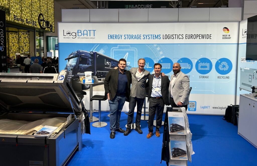 Vier Männer lächeln in einem Messestand mit blauer Rückwand und Werbegrafik für Logistiksysteme.