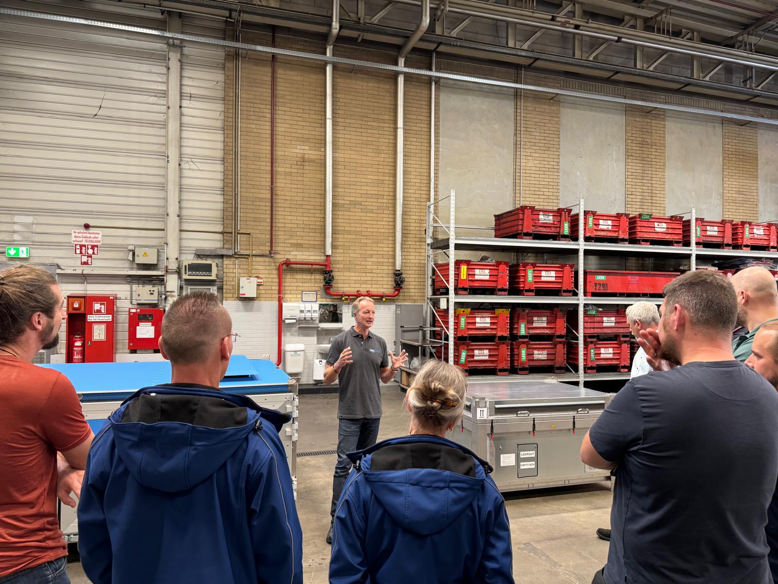 Eine Gruppe von Menschen hört einem Mann in einer Lagerhalle bei einem Vortrag zu.