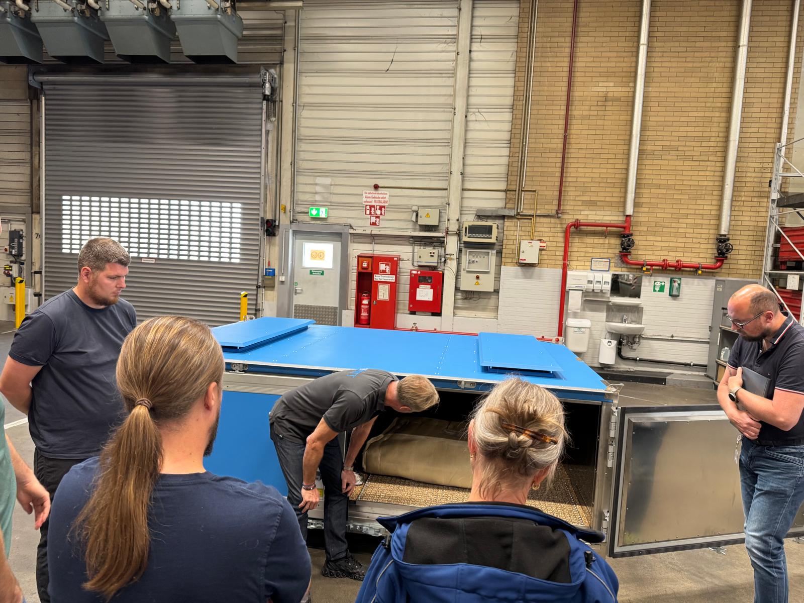 Ein Mann untersucht einen großen blauen Metallbehälter, umgeben von Zuschauern in einer Lagerhalle.