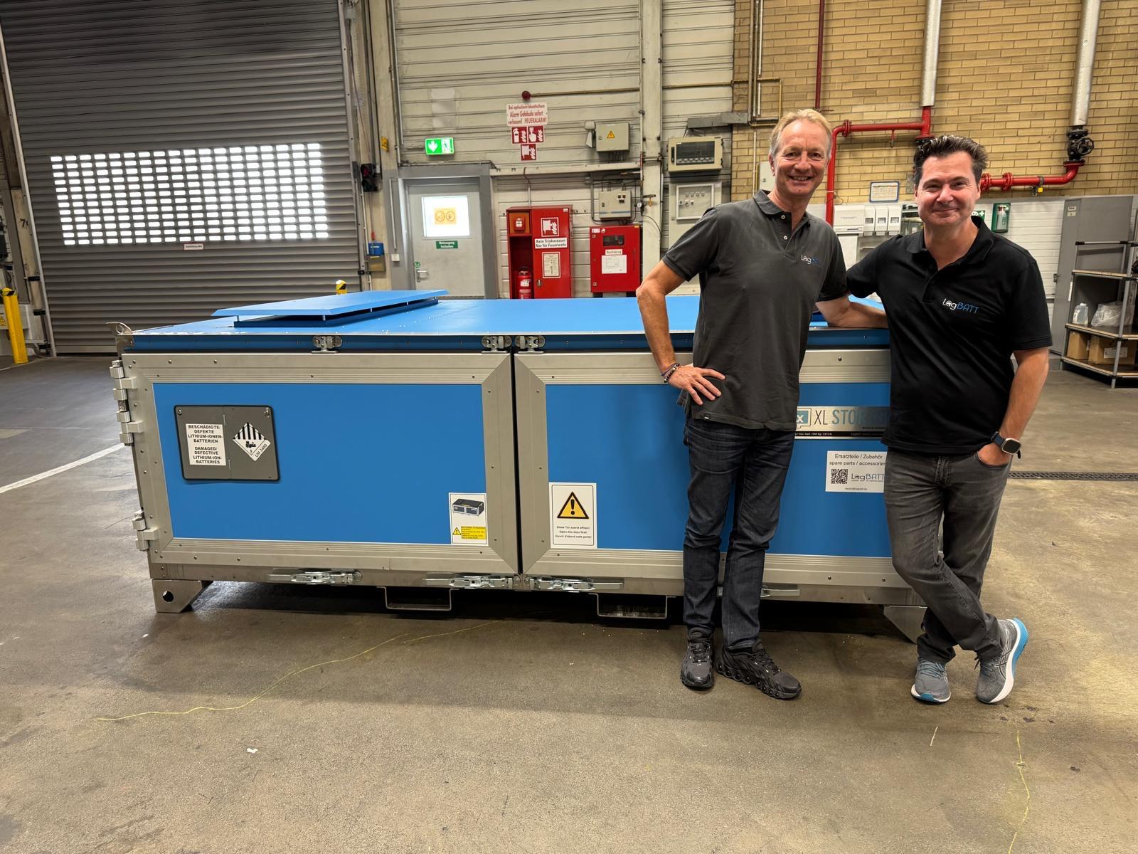 Zwei lächelnde Männer in schwarzen Polohemden lehnen an einer großen blauen Kiste.