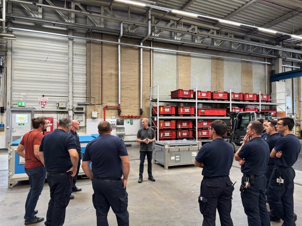 Eine Gruppe von Männern in Arbeitskleidung steht in einer Fabrikhalle und hört einem Mann aufmerksam zu.