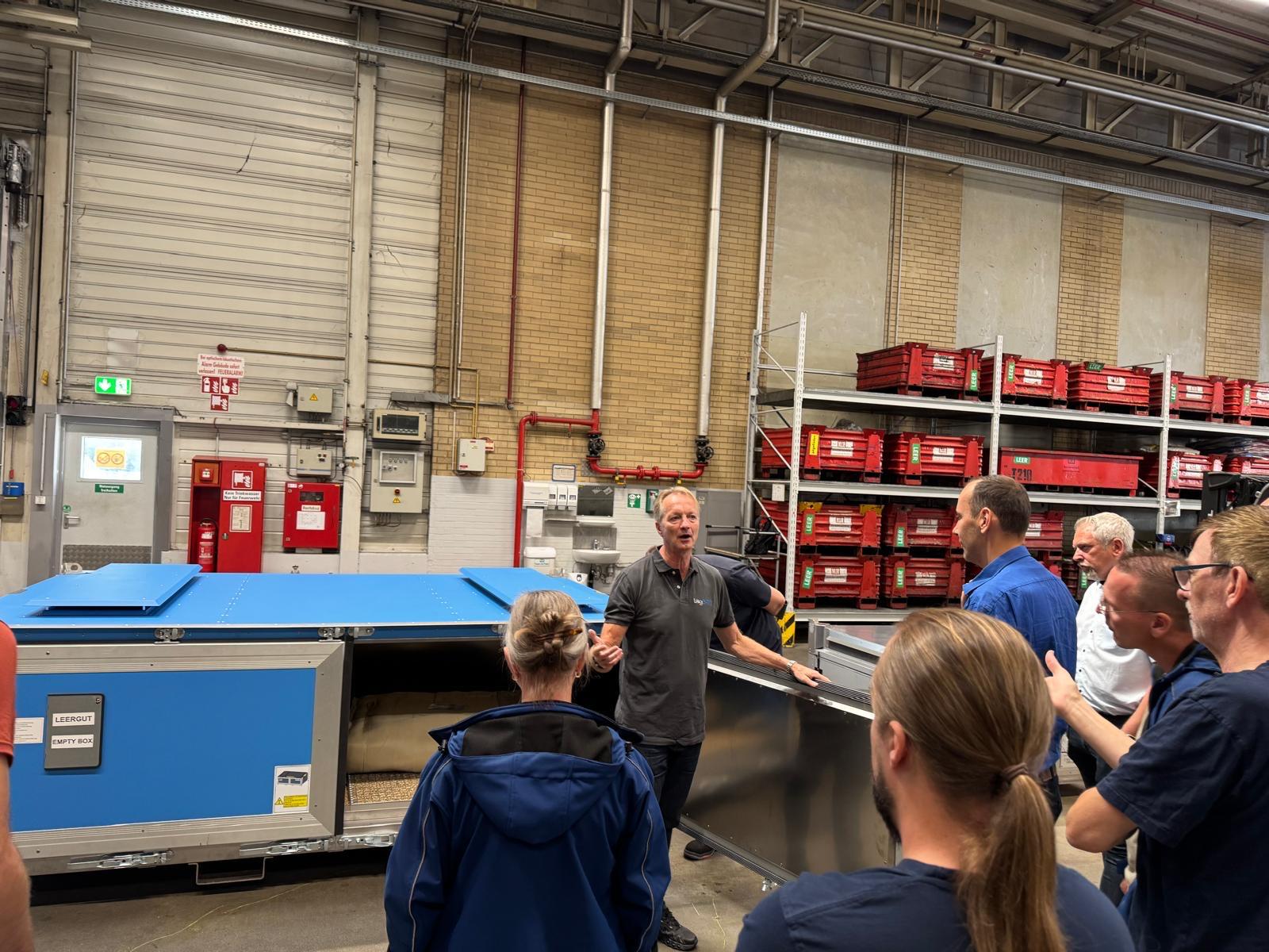 Ein Mann erklärt einer Gruppe Menschen eine große blaue Maschine in einer Lagerhalle.