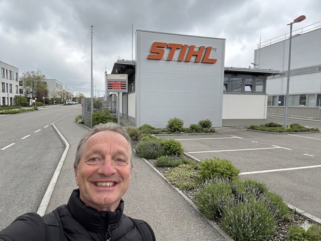 A person with a neutral expression is standing in front of a grey STIHL building under a cloudy sky