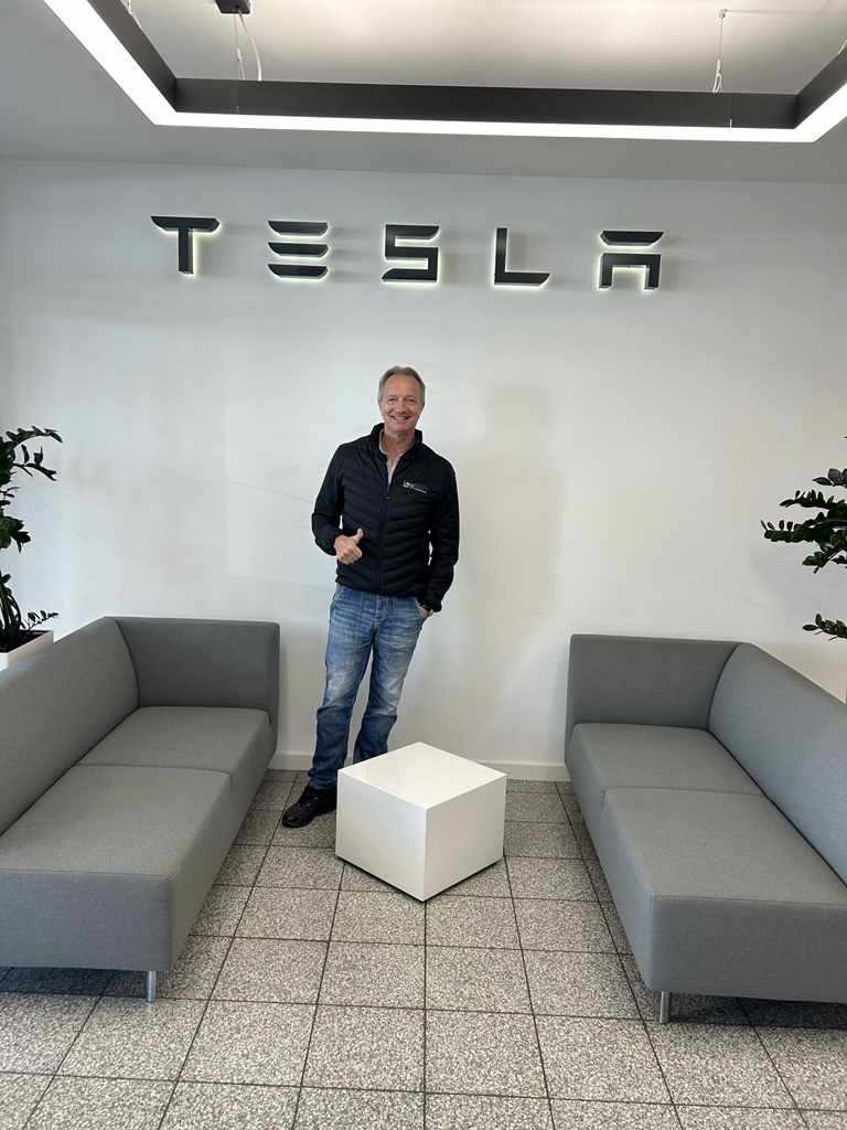A man wearing jeans and a black jacket is smiling in front of the Tesla logo and giving a thumbs-up
